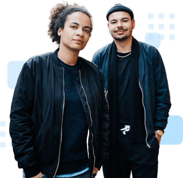 Two confident individuals standing side by side and wearing black jackets; one with curly hair and the other in a beanie.