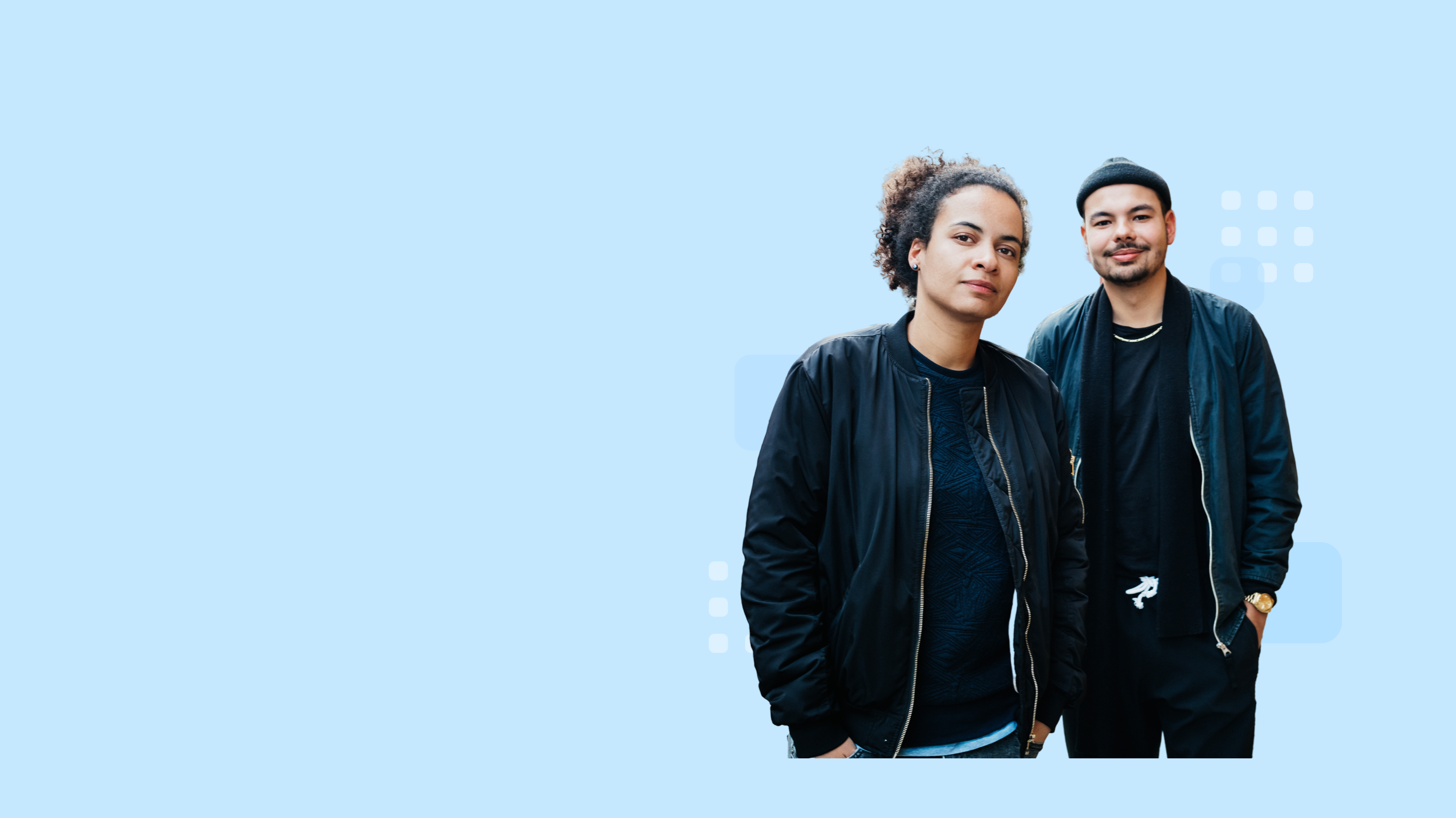 Two confident individuals standing side by side and wearing black jackets; one with curly hair and the other in a beanie.