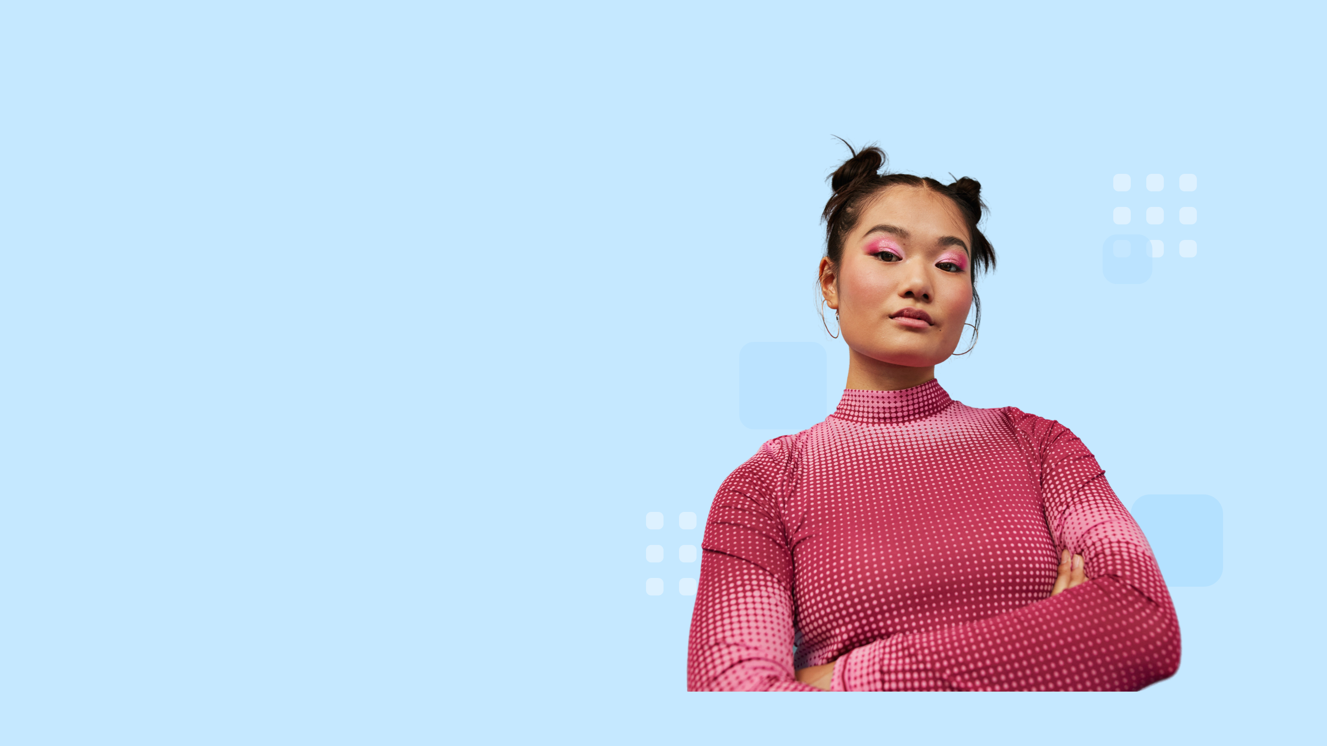 Confident individual with styled hair in space buns, wearing a pink patterned top, and standing with arms crossed.